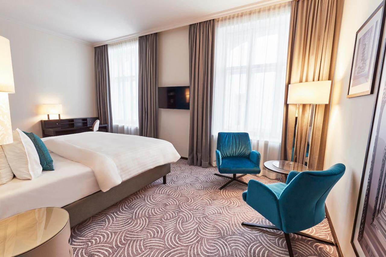 Steigenberger Hotel Herrenhof Vienna Esterno foto The photo shows a well-decorated hotel room. It features a large bed with soft bedding and modern furnishings. There are two large windows with sheer curtains that allow natural light to enter. A flat-screen TV is mounted on the wall across from the 