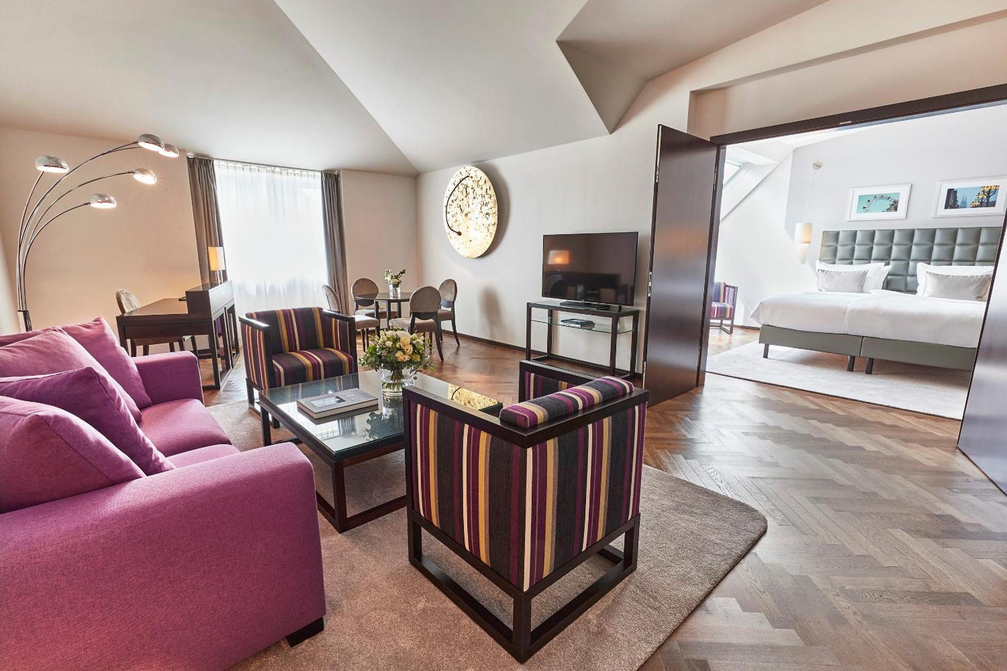 Steigenberger Hotel Herrenhof Vienna Esterno foto The photo shows a modern, stylish living space that appears to be part of a hotel suite. The room features a cozy seating area with a purple sofa and two striped chairs around a glass coffee table. A flat-screen TV is mounted on the wall. Large windo